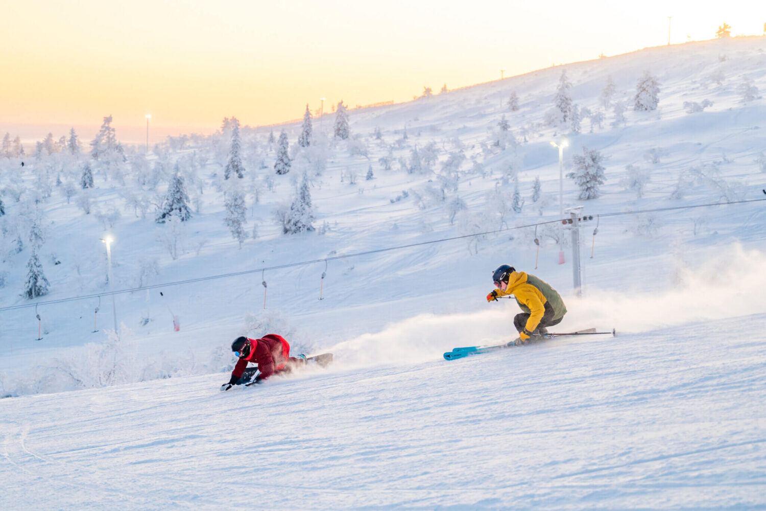 Two skiers carving down a sunlit slope, snow spray rising; frosted trees and a lift line in the background.