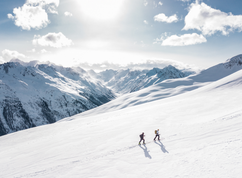 Two ski tourers crossing a vast snow-covered mountain slope beneath a blue sky.