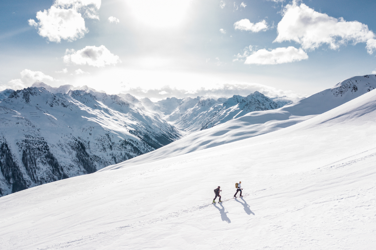 Two ski tourers crossing a vast snow-covered mountain slope beneath a blue sky.